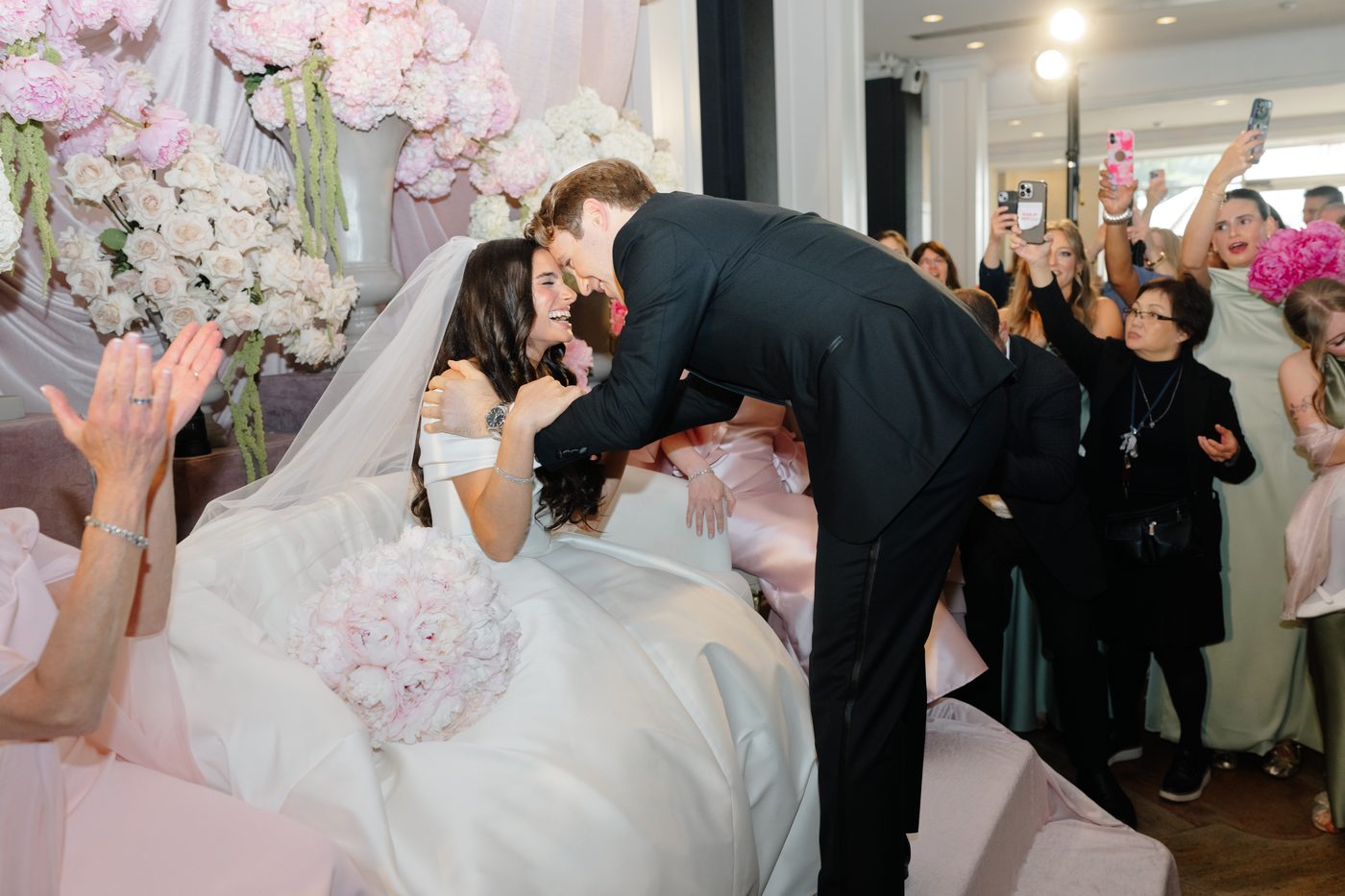 The groom leans in to the bride at the bedeken, surrounded by family and bridesmaids in sage green and blush.