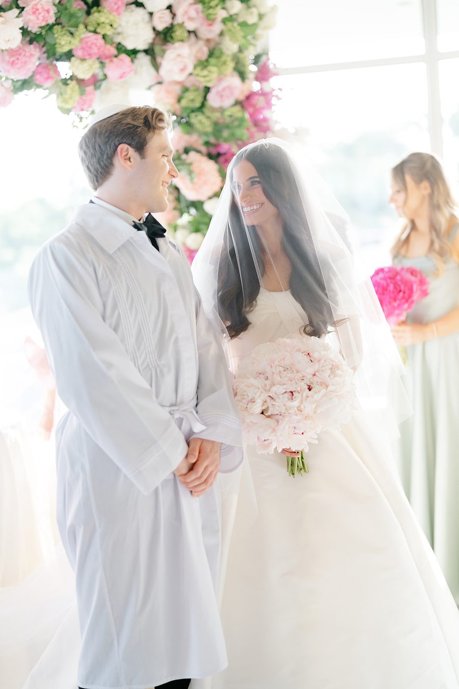 The groom in his white kittel stands beside the newly veiled bride, who holds her peony bouquet.
