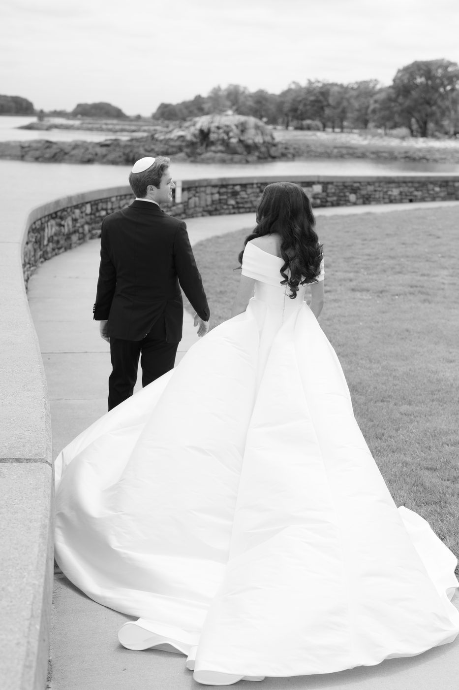 Black and white photo of the couple walking the curved seawall path, the bride's long train sweeping behind her.