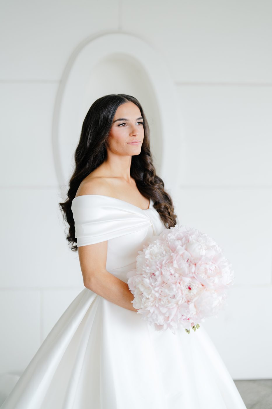 Bridal portrait in an off-the-shoulder ivory gown holding a full bouquet of blush peonies.