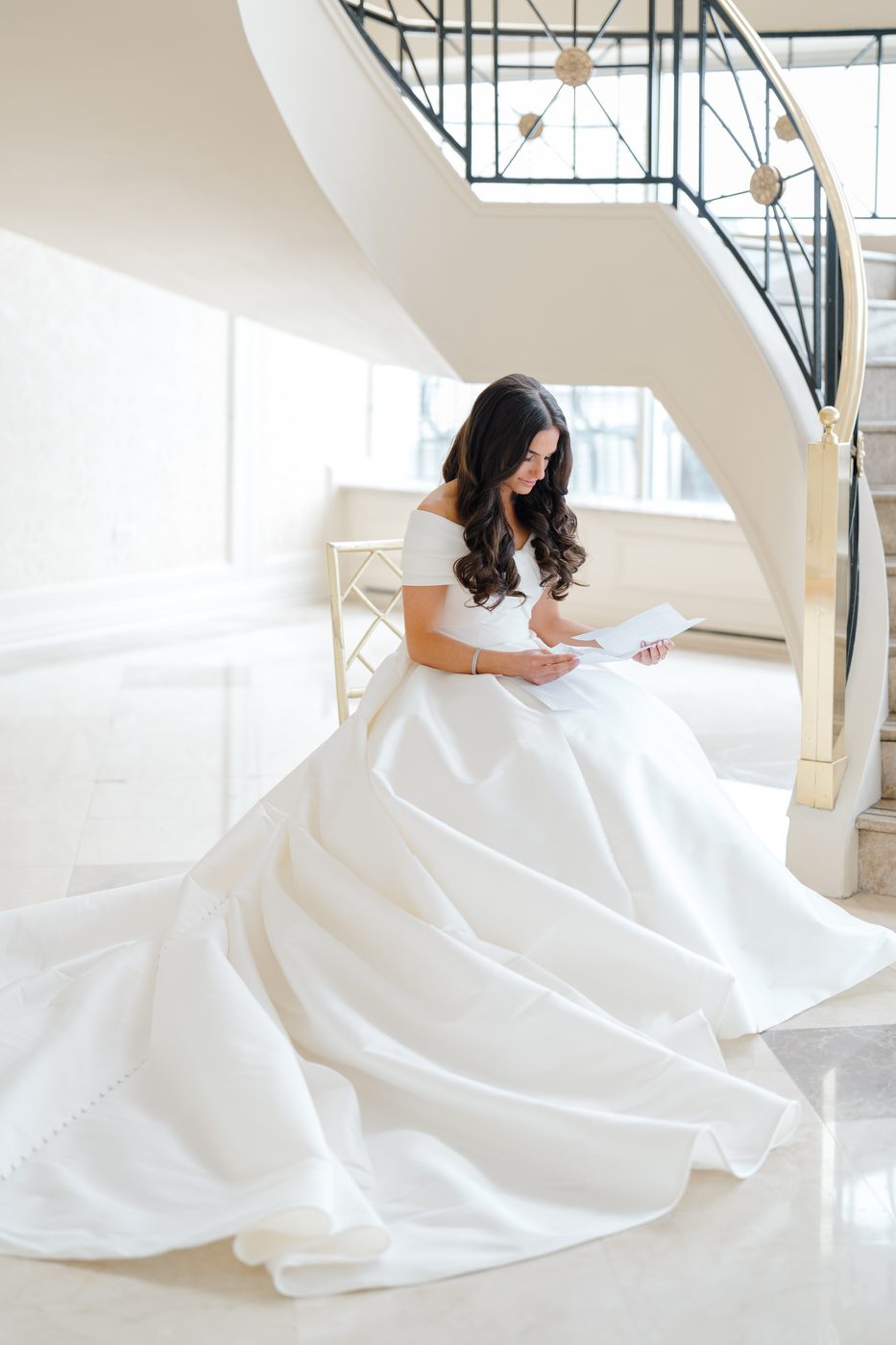 The bride sits at the foot of the curved staircase at Glen Island Harbour Club reading a letter from the groom, her ivory ball gown spread across the marble.