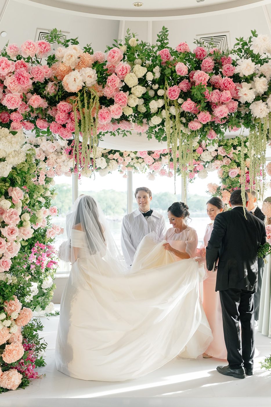 The full ceremony beneath the floral chuppah with guests filling the windowed room.
