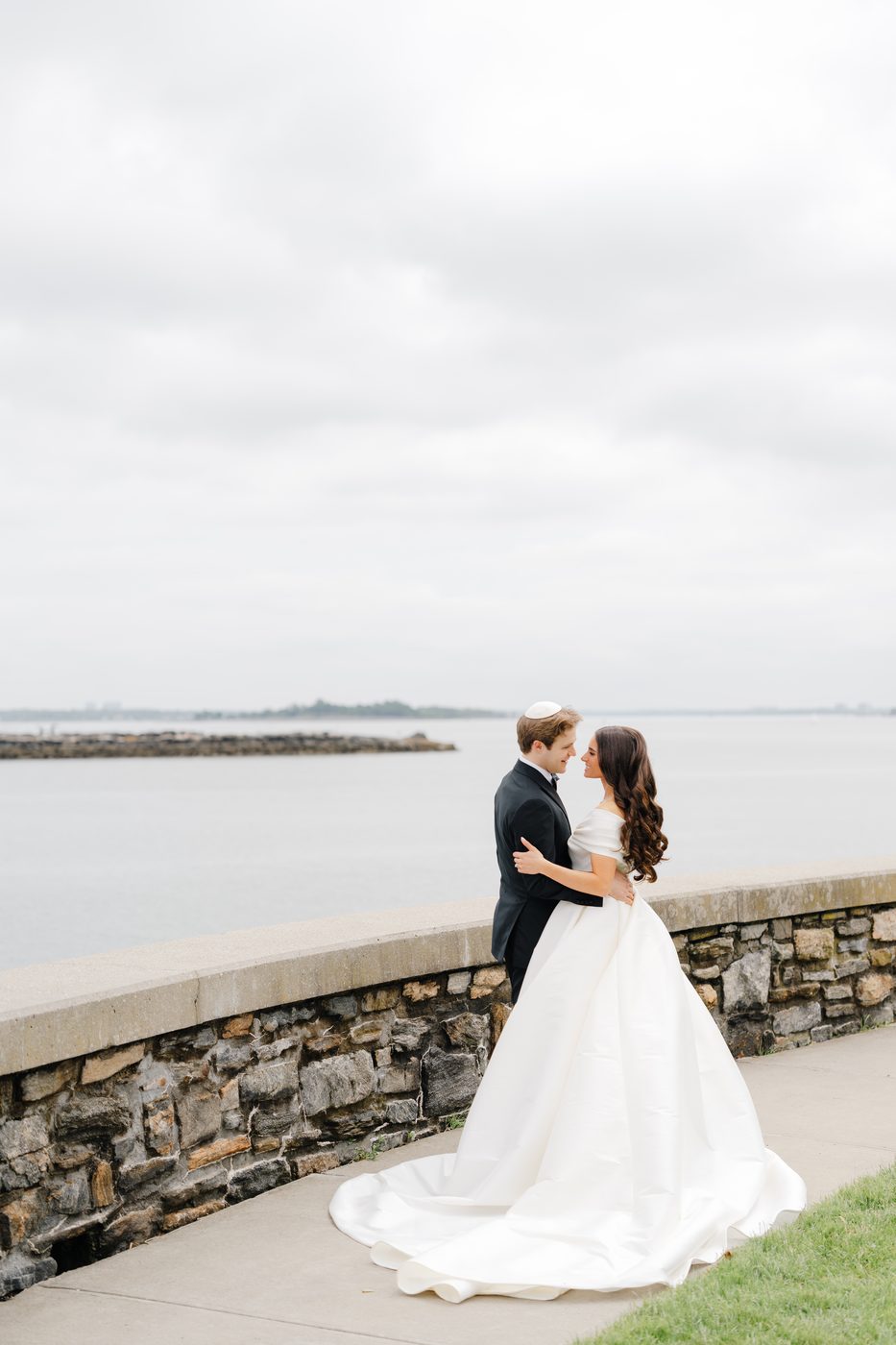 The bride and groom stand forehead to forehead along the stone seawall with Long Island Sound behind them.
