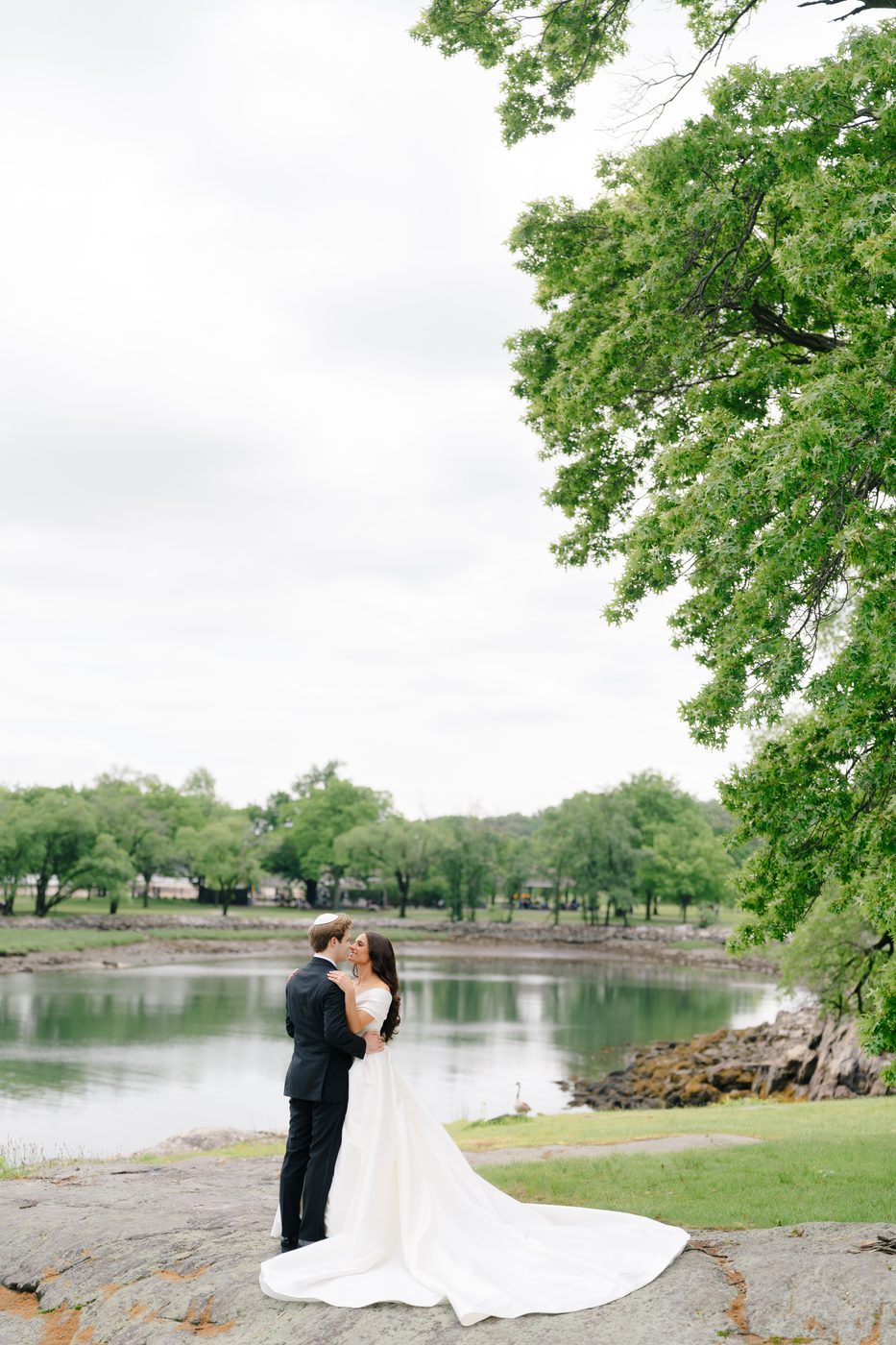 The couple kiss on the rocks above the lagoon at Glen Island, framed by a canopy of green.