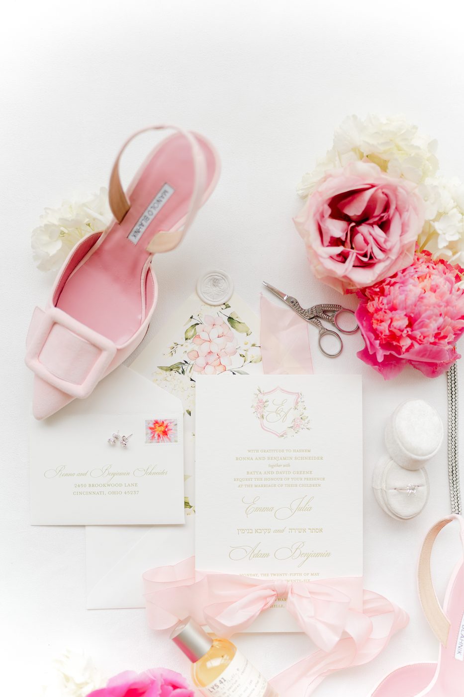 Flat lay of the wedding invitation suite with gold Hebrew and English lettering, blush peonies, pink buckle heels, and the rings.