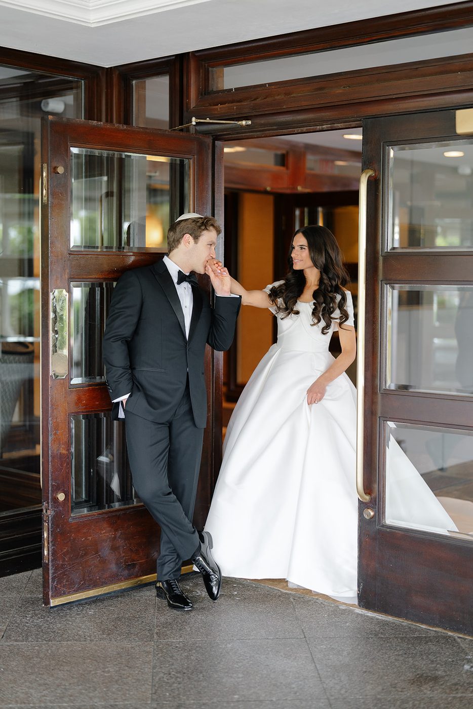 The groom kisses the bride's hand in the doorway of the club, her train sweeping the threshold.