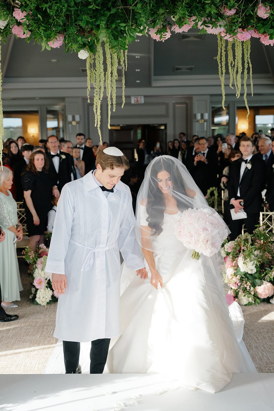 The groom in his kittel walks with the veiled bride beneath hanging greenery and blooms.