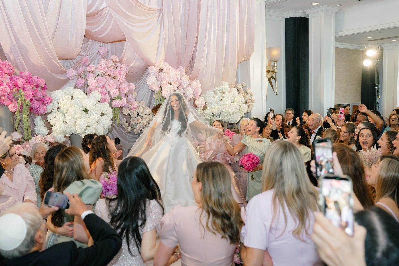 The bride is seated beneath blush draping and banks of pink and white peonies during the kabbalat panim as guests dance around her.