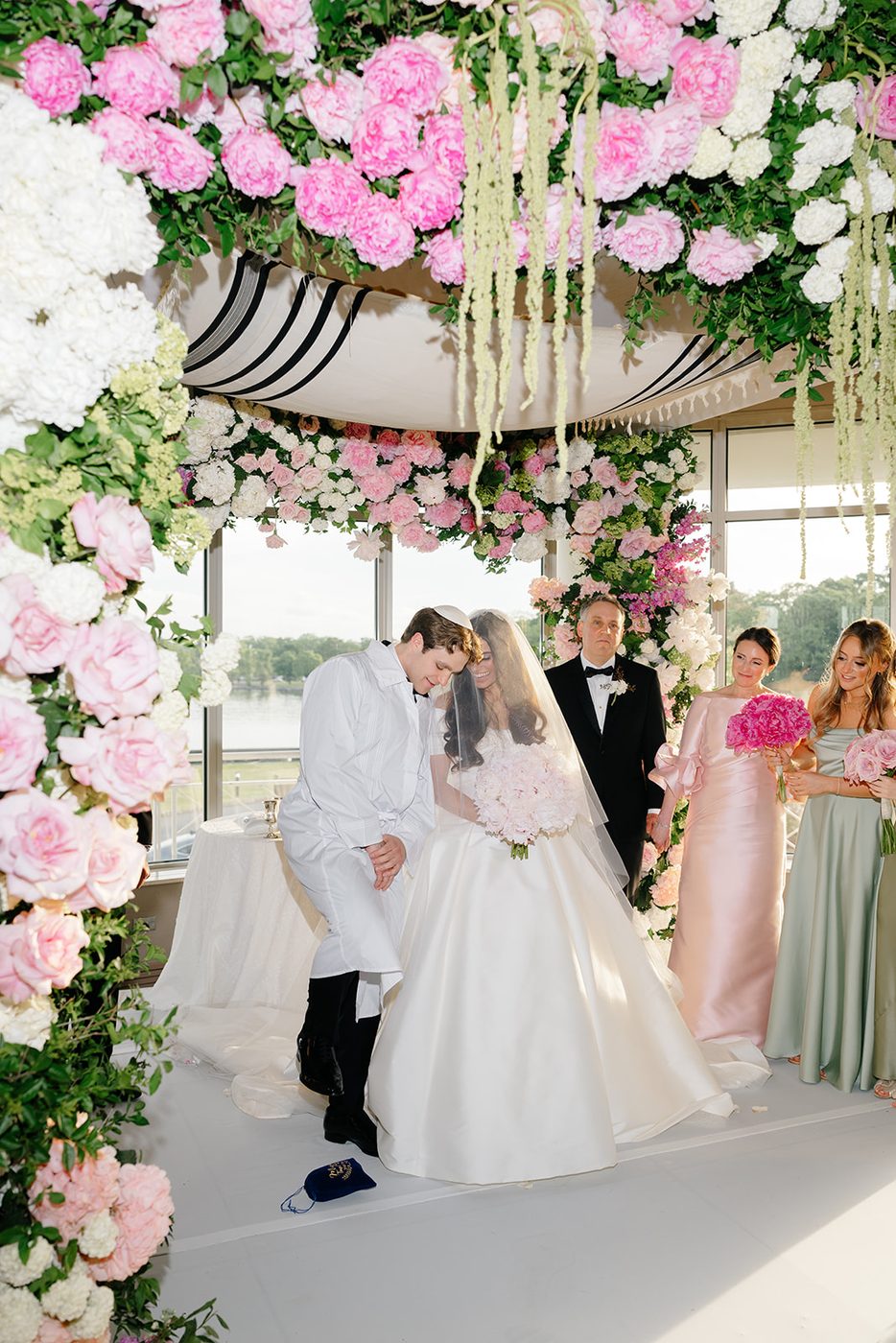The bride and groom kiss beneath the pink floral chuppah as guests look on.