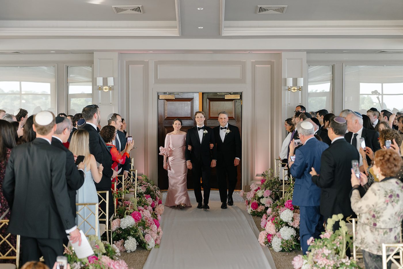Guests rise as the processional begins down the white aisle toward the floral chuppah.