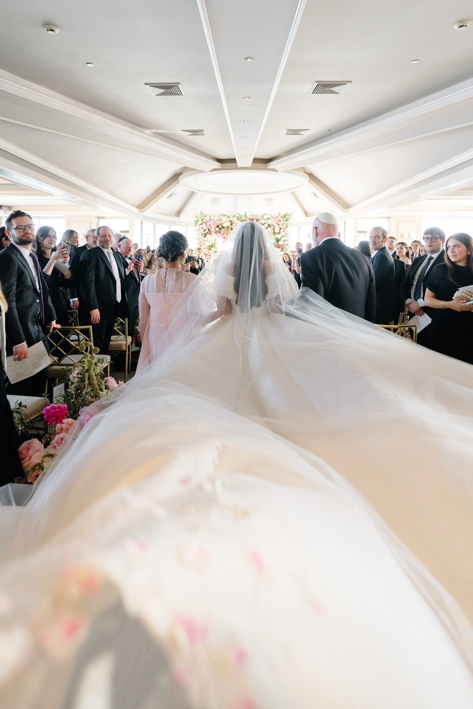 The bride walks the aisle with her veil sweeping behind her toward the pink and white floral chuppah.