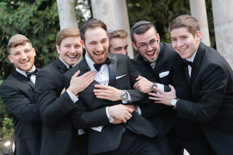 The groom laughs as his groomsmen wrap him in a group hug in front of the stone columns.