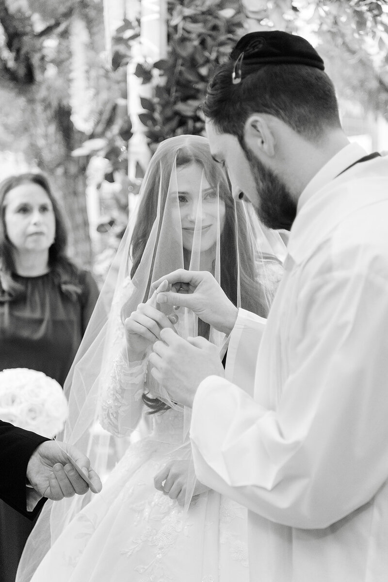 Black and white photo of the groom lowering the veil over the smiling bride during the badeken.