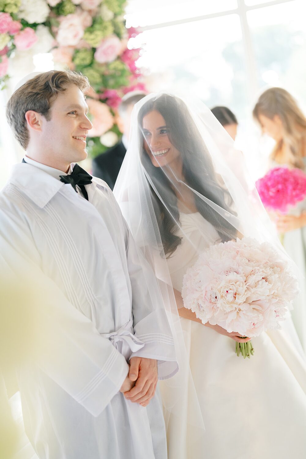 The groom smiles at his veiled bride during the bedeken, her peony bouquet in hand.