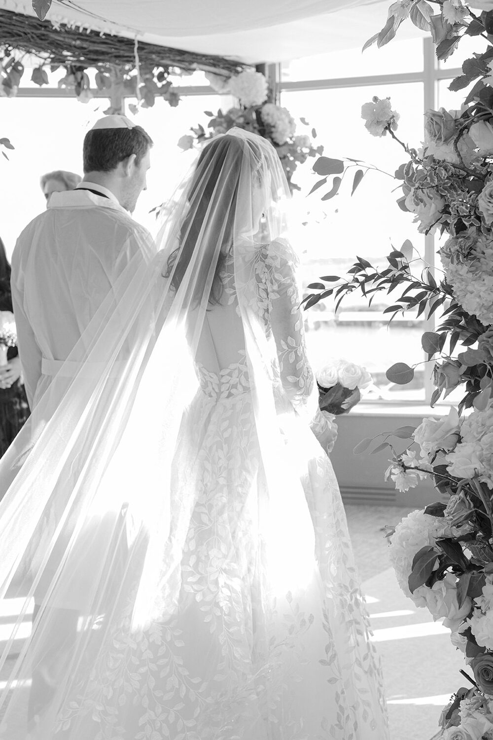 Seen from behind in black and white, the veiled bride stands beside the groom under the chuppah.