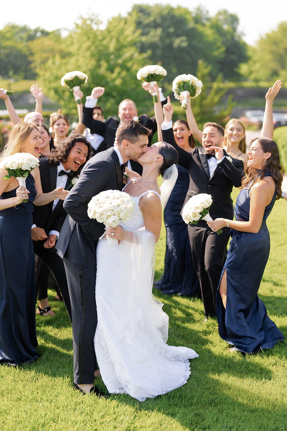 The bridal party cheers with bouquets raised as the couple kisses on the lawn.