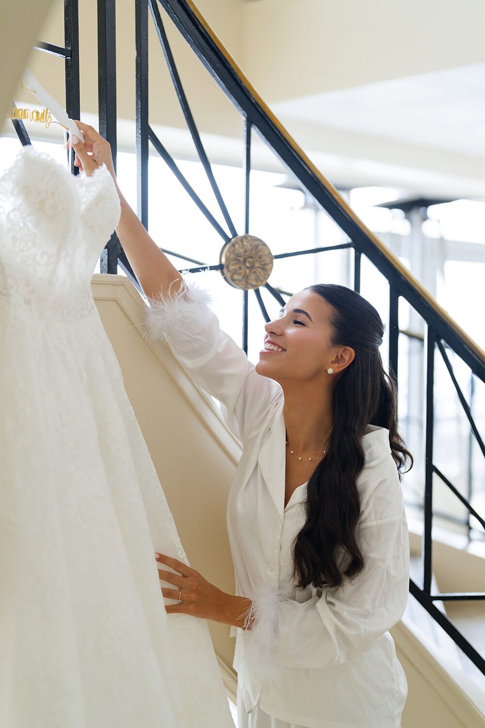 Bride reaches for her gown hanging from the staircase railing while getting ready at Glen Island Harbour Club.