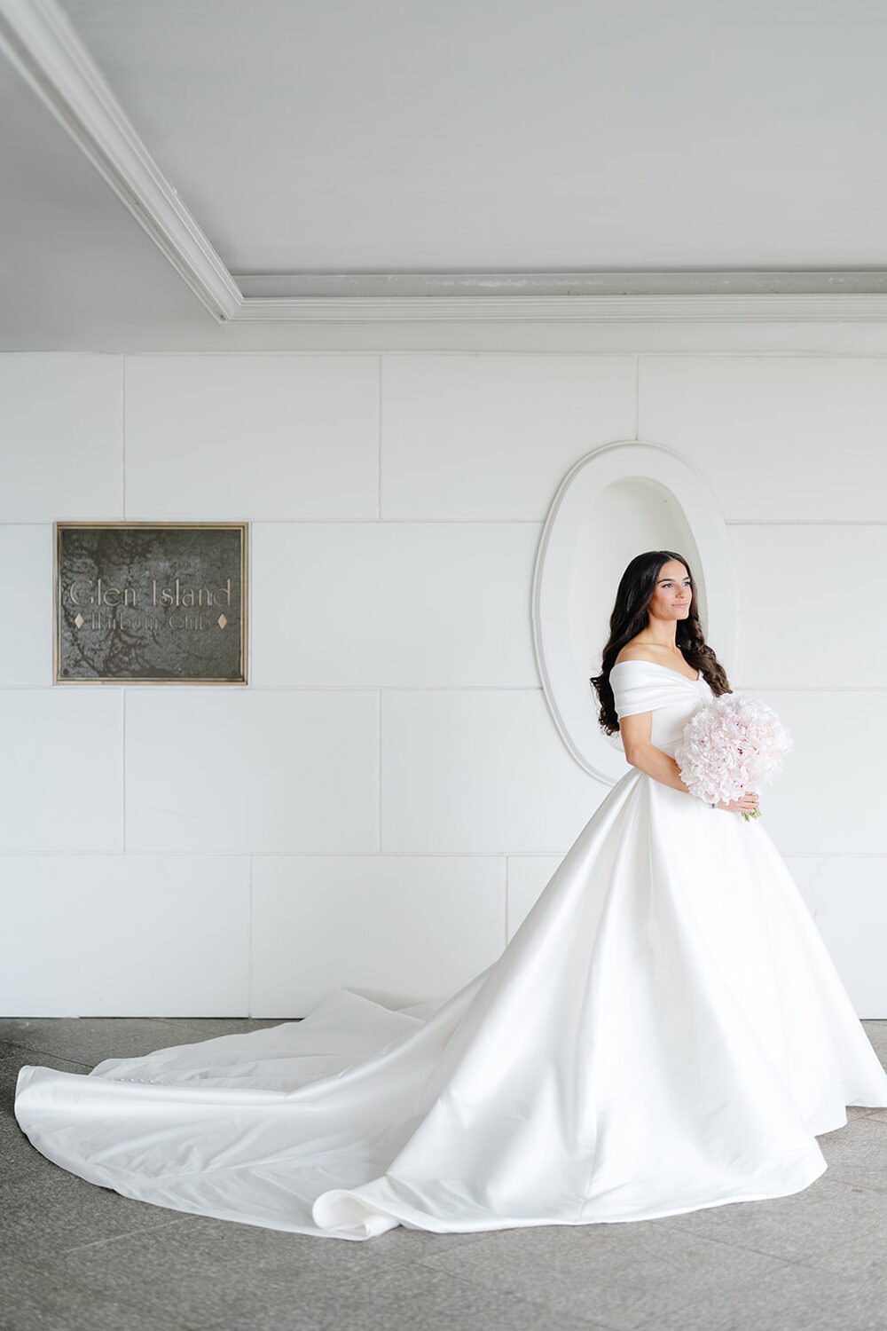 The bride stands before the oval mirror in the mansion's white marble hallway, train pooled behind her.