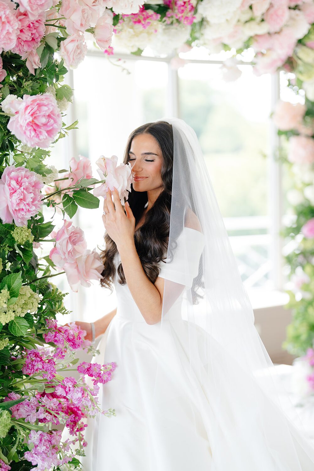 The bride pauses to smell pink peonies woven into the floral arch.