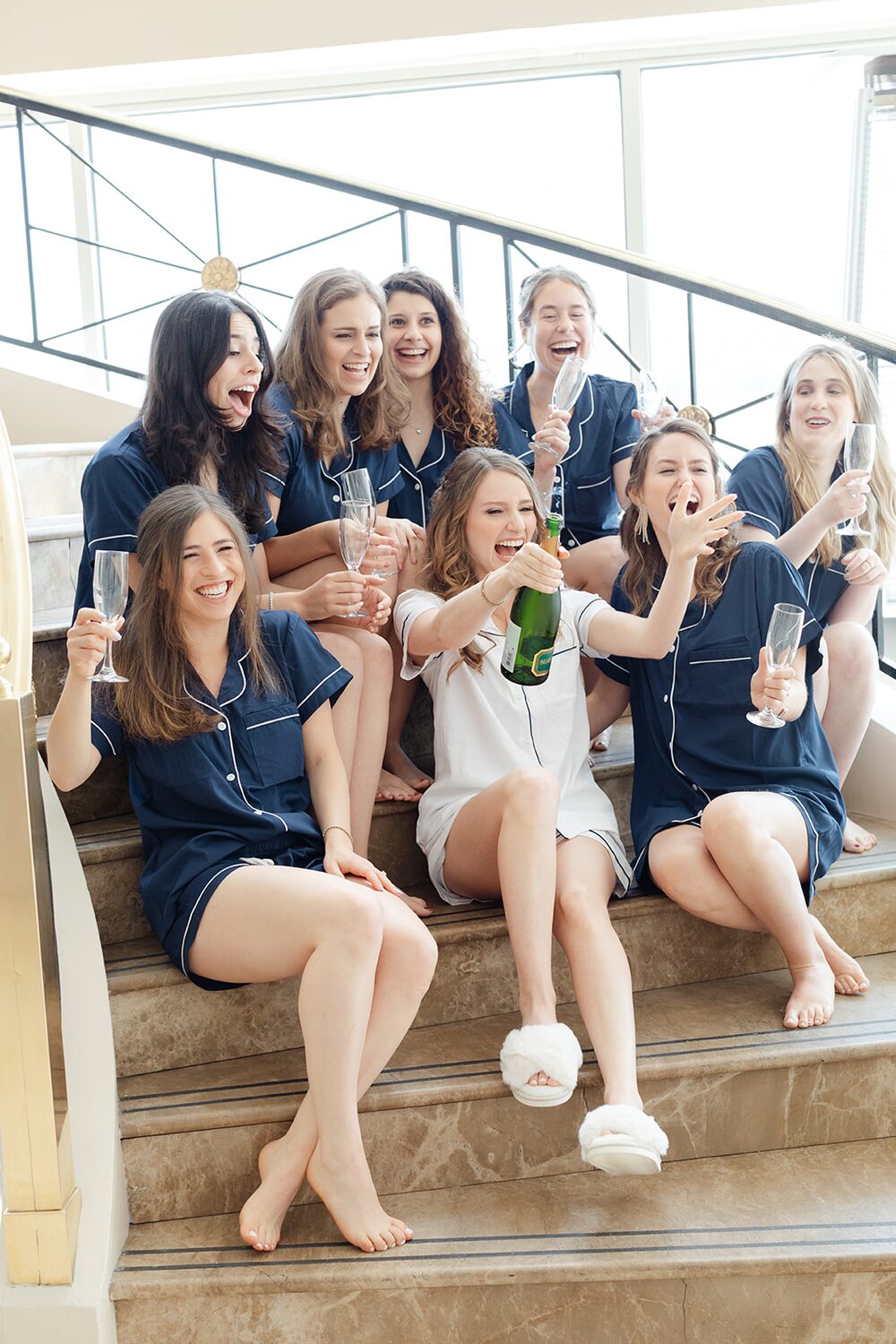 Bridesmaids in navy pajamas pop champagne on the staircase while getting ready.