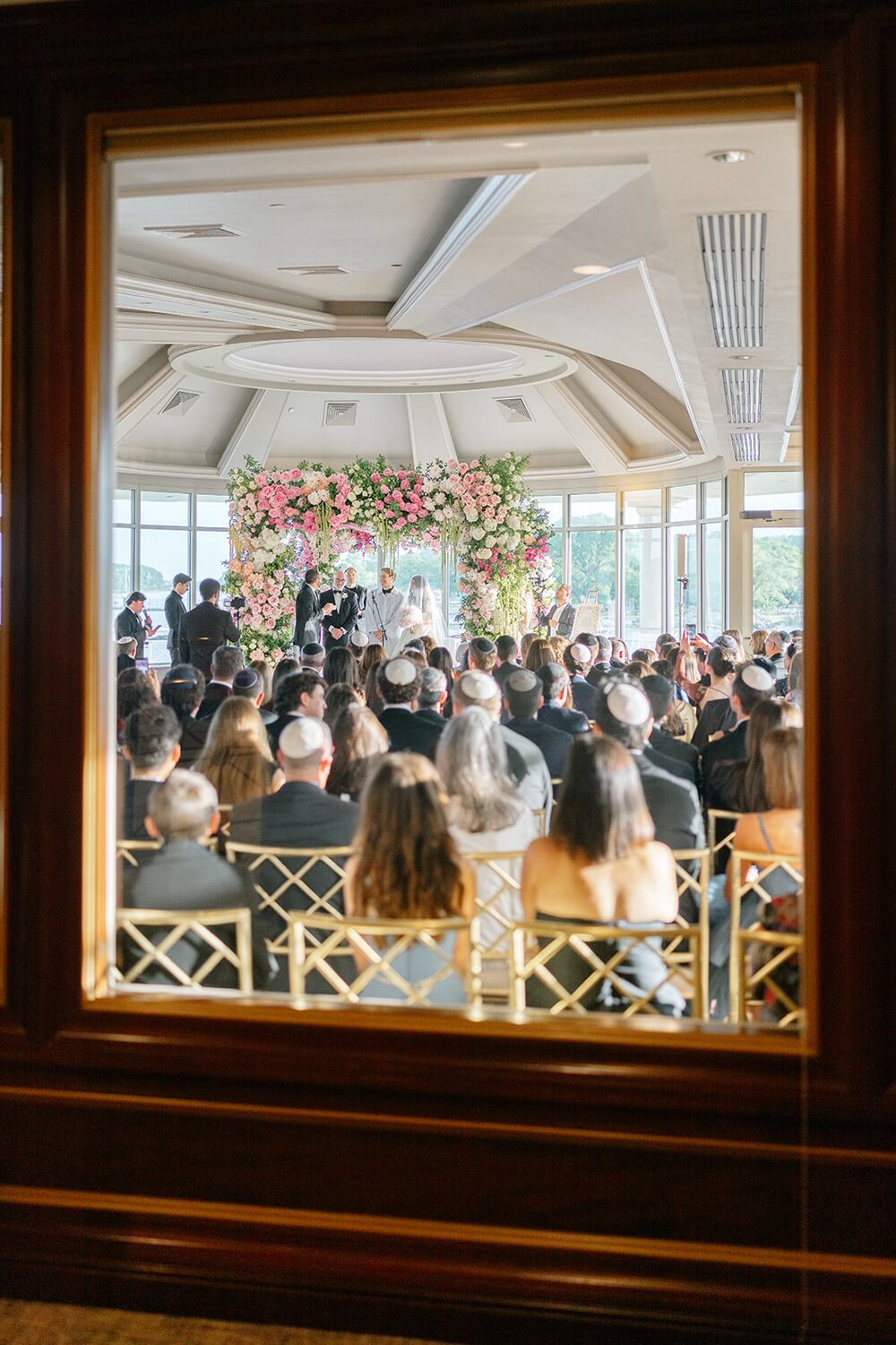 The chuppah ceremony glimpsed through a framed doorway, guests filling the glass room.