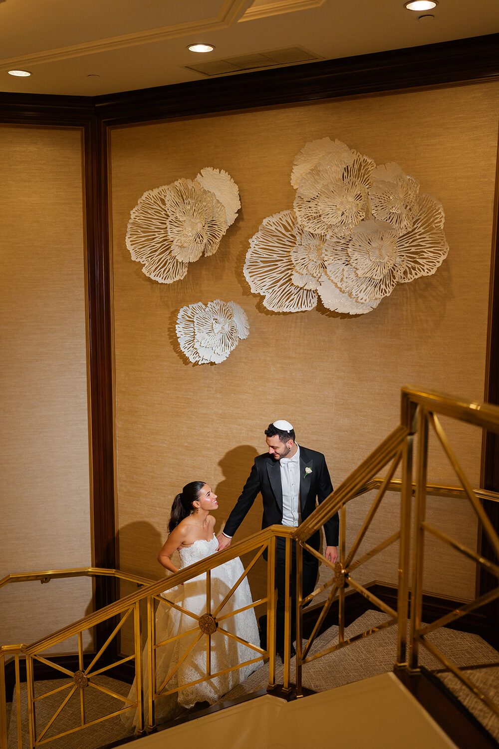 Bride and groom descend the gold-railed staircase beneath sculptural coral wall art.