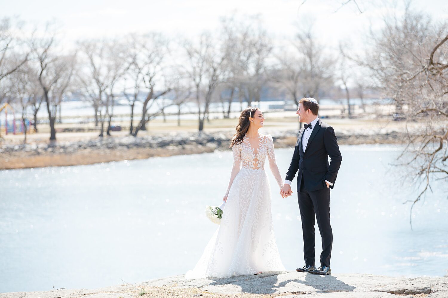 The couple stands at the pond's edge in early spring, bare trees mirrored in the water.