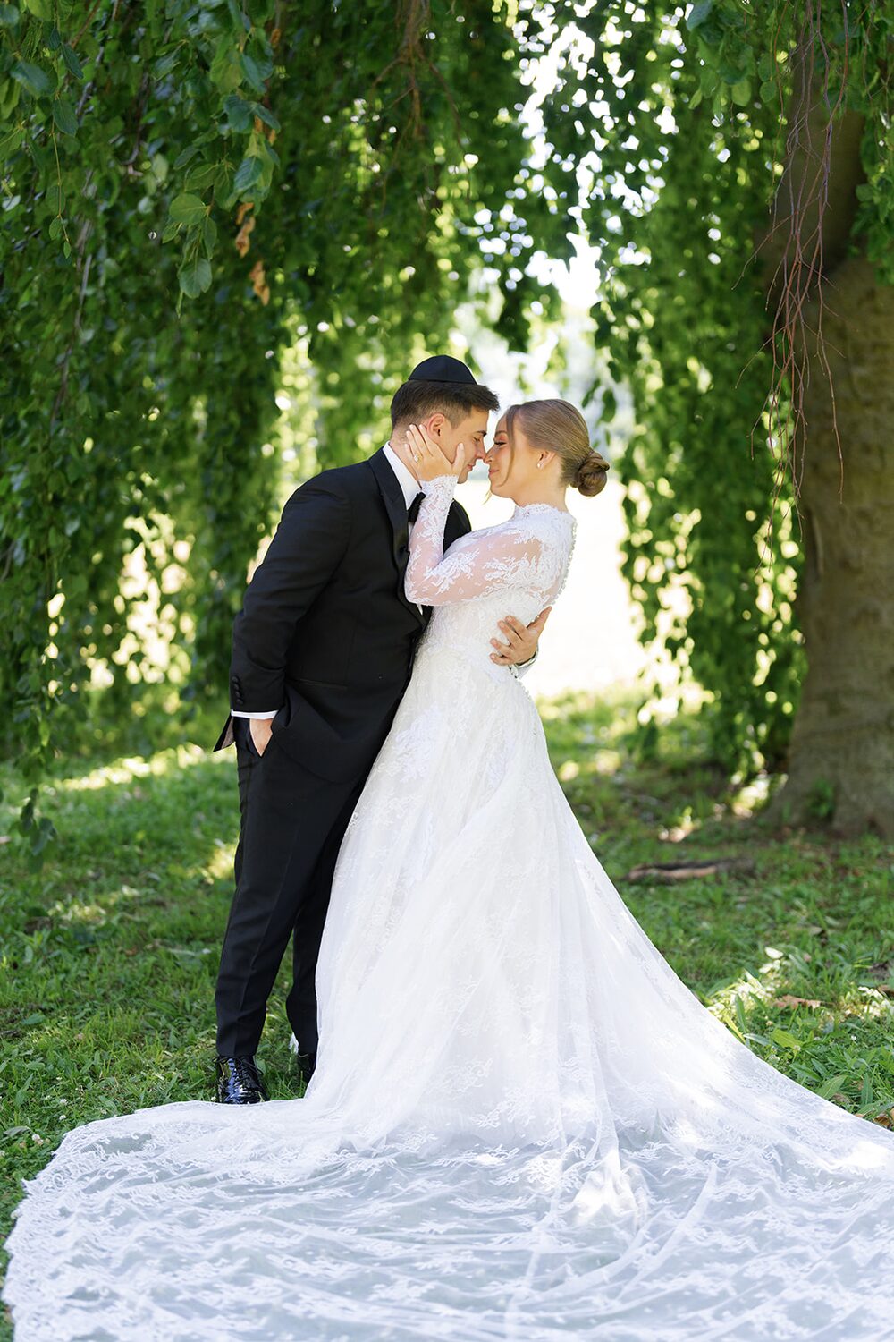 Eliana and Elan embrace beneath a willow on Glen Island, her lace train spread across the grass.