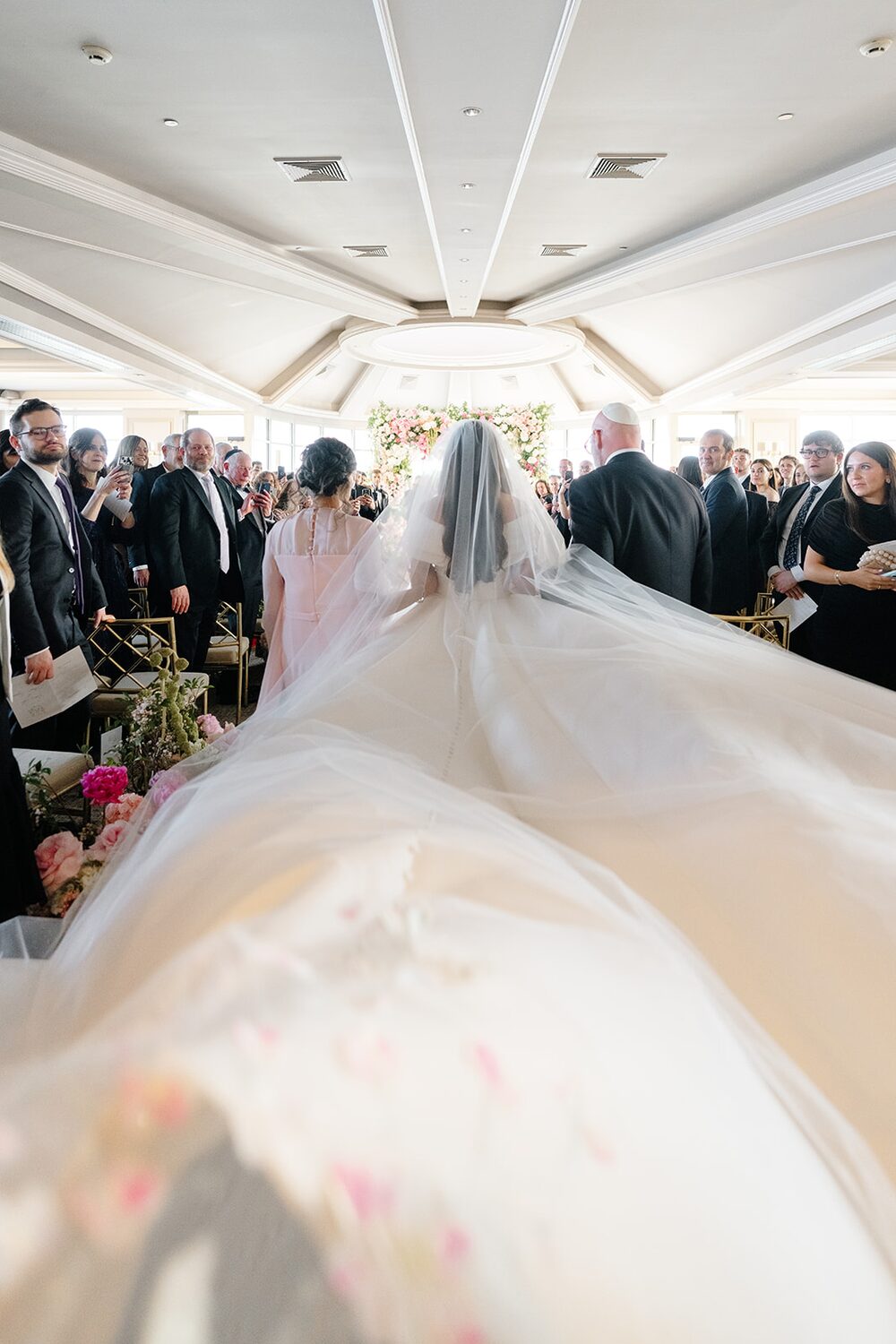 Emma's cathedral veil fills the aisle as she and Adam stand before the chuppah.