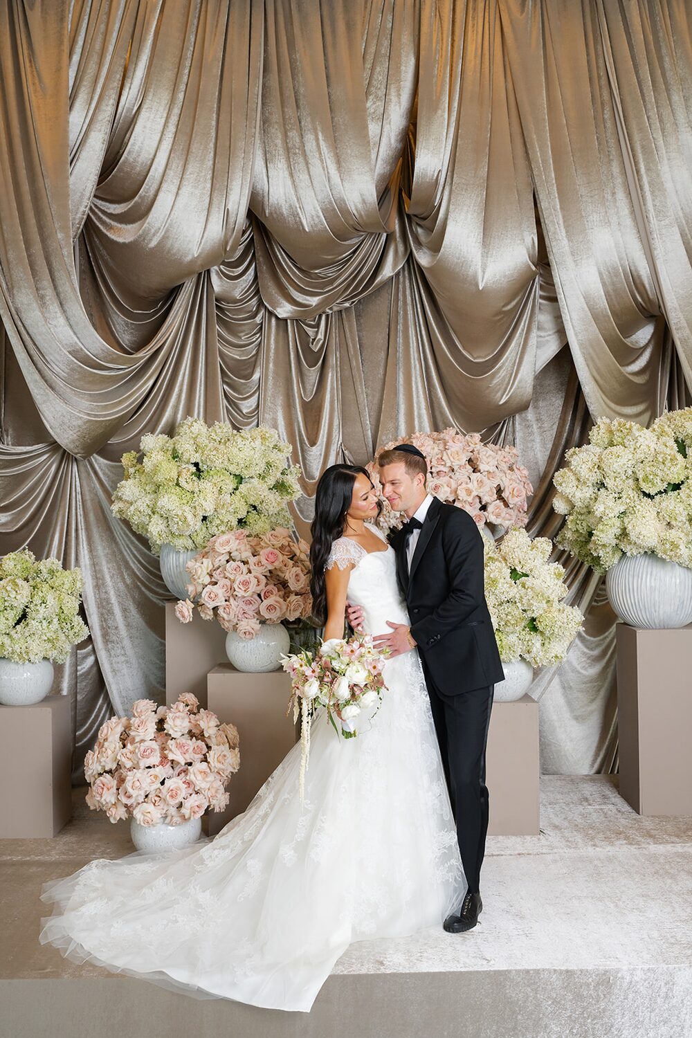 The couple poses before the gold drapery and towering hydrangea arrangements of the badeken stage.