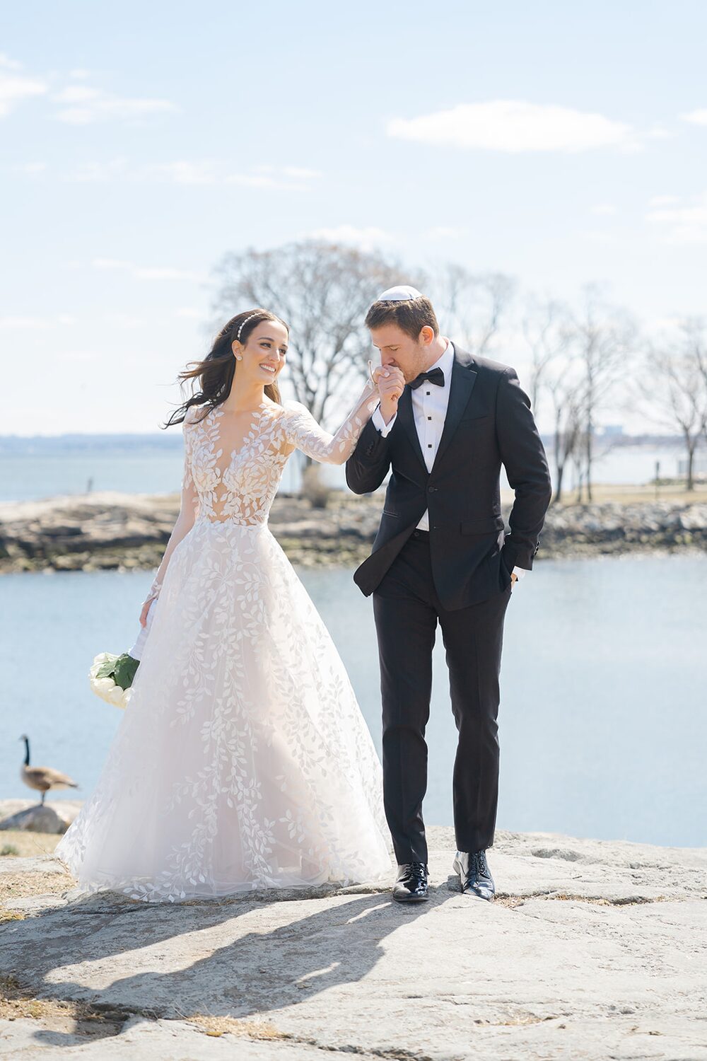 The groom kisses the bride's hand on the rocky shoreline of Long Island Sound.