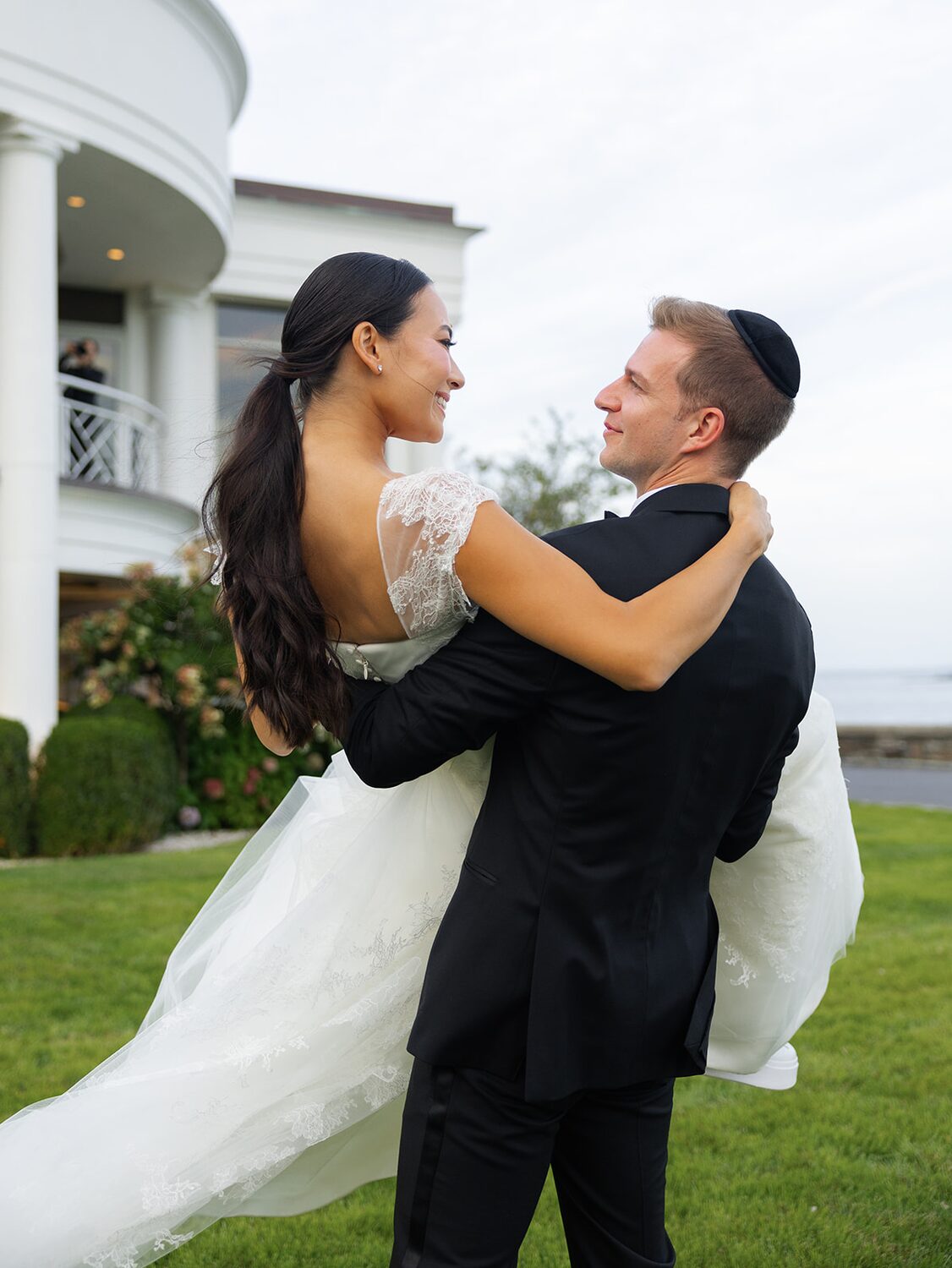 The groom lifts his bride on the mansion lawn beneath the white balconies.