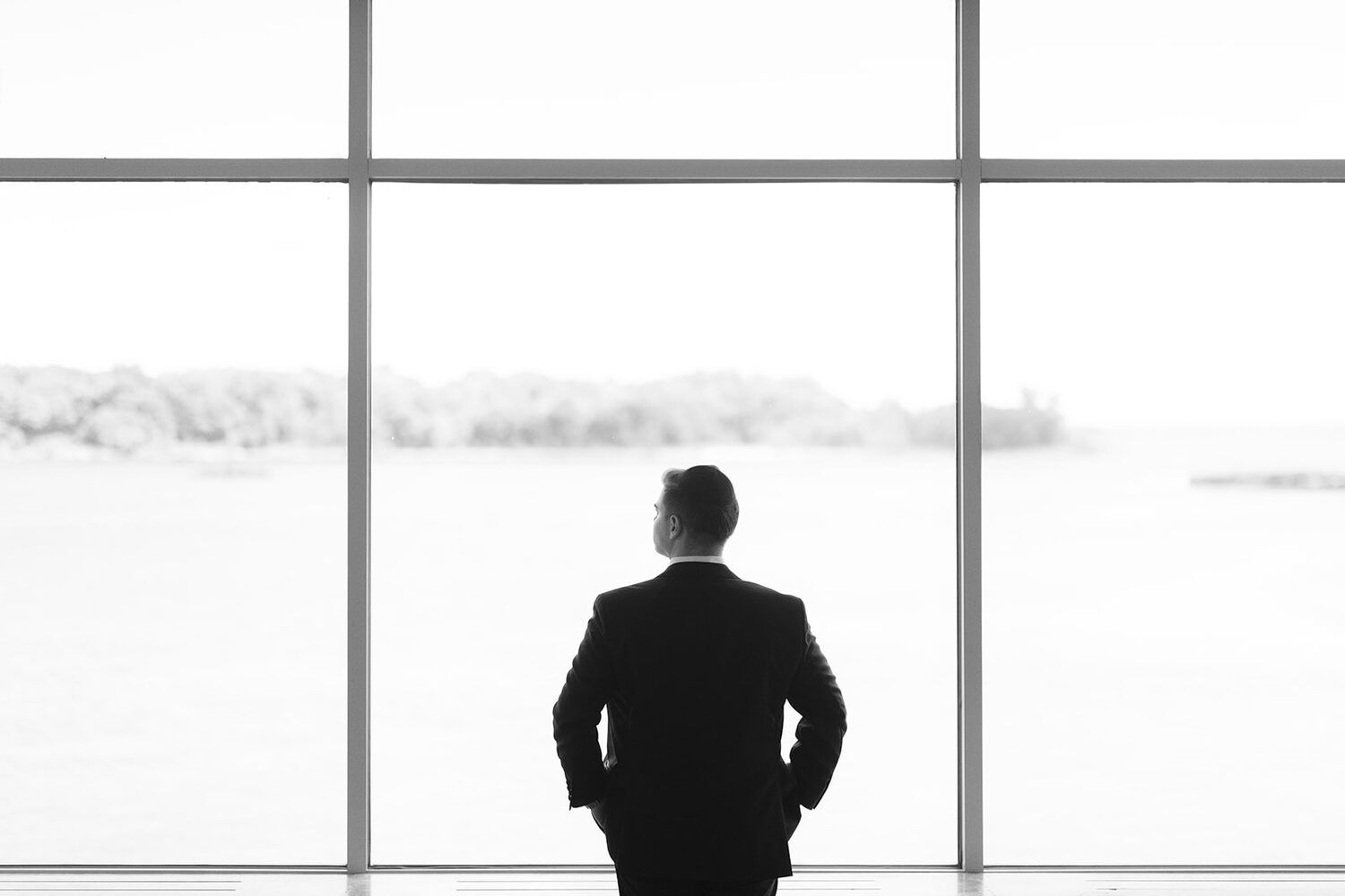 The groom stands in silhouette at the floor-to-ceiling windows overlooking the water.