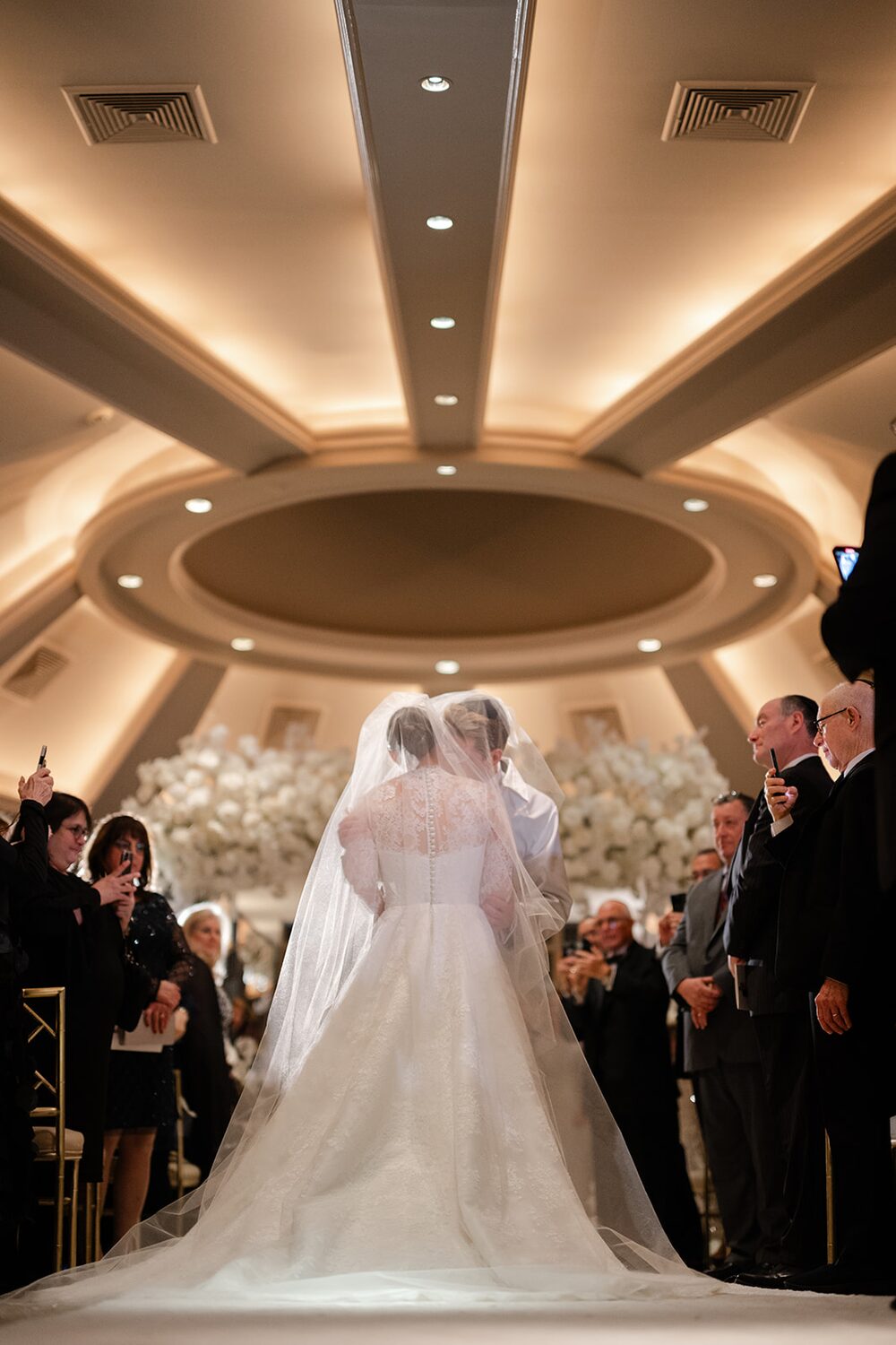 Hannah walks the aisle toward Philip and the white floral chuppah as guests rise.