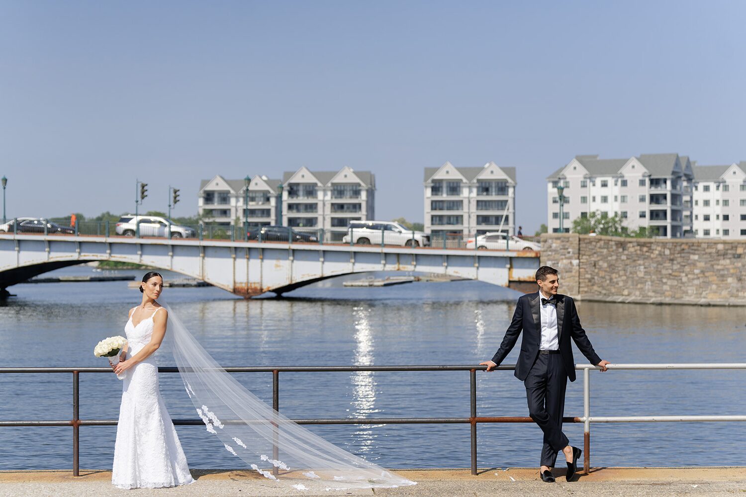The bride and groom lean on the harbor railing with the New Rochelle bridge beyond.