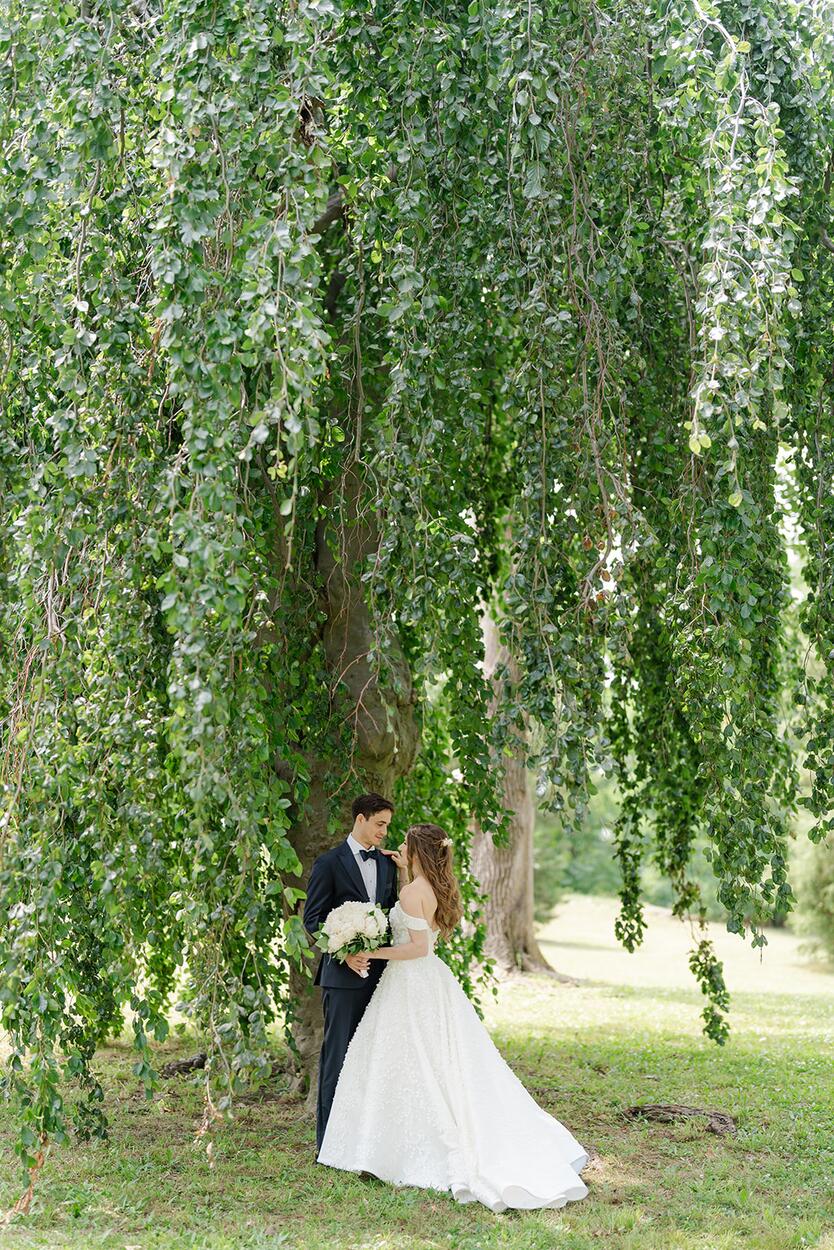 The couple pauses under a tall June willow, bouquet of white blooms in hand.