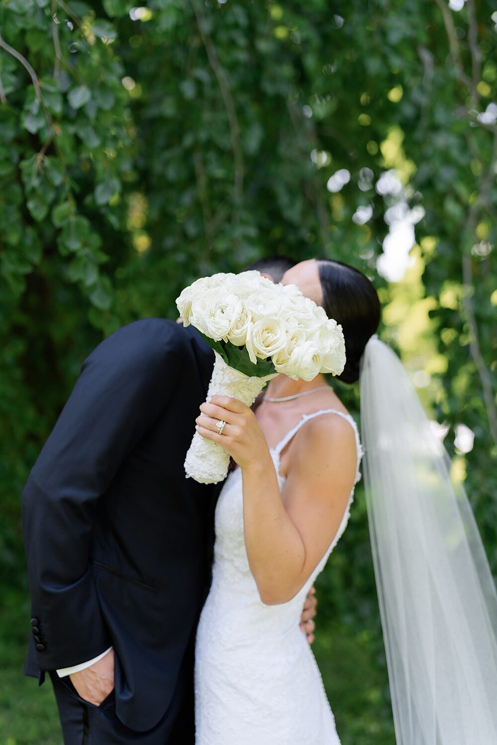 The groom leans in for a kiss behind the bride's white rose bouquet.