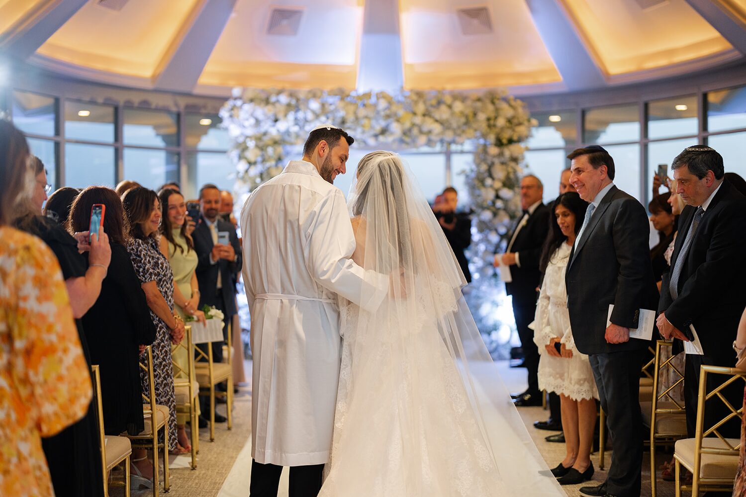 The groom in a traditional white kittel stands with his veiled bride under the chuppah as guests look on.