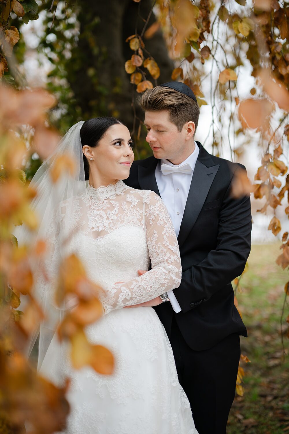 In long lace sleeves, the bride leans into the groom among golden autumn leaves.