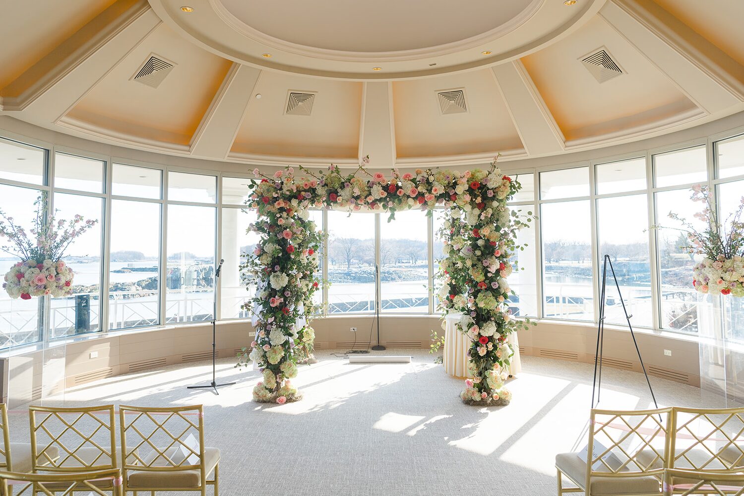 A rose-covered chuppah stands ready in the solarium, water views wrapping the room.
