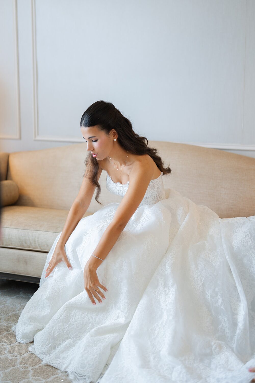 The bride arranges her lace gown while seated on a cream sofa before the ceremony.