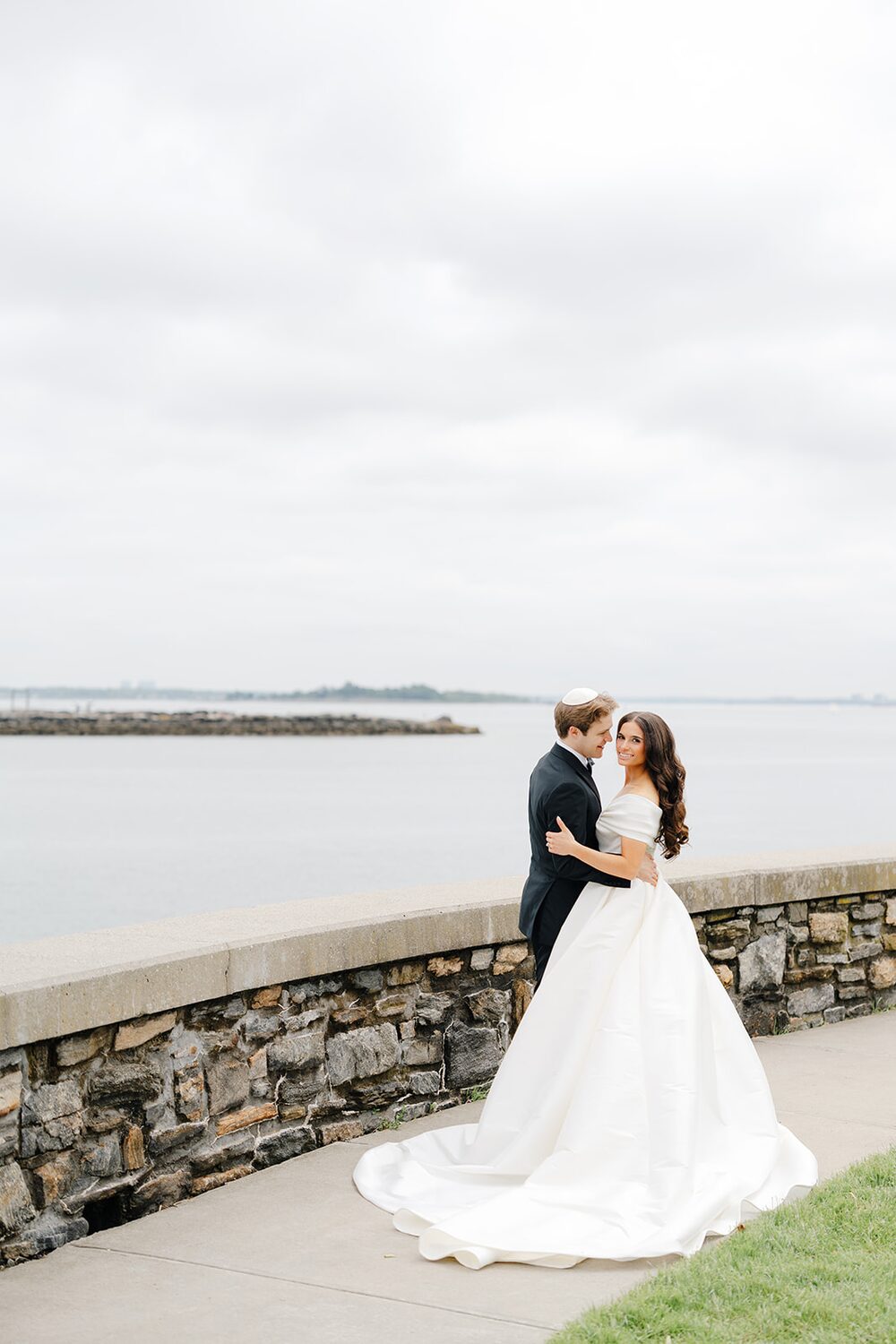 The couple embraces at the seawall under soft overcast light on Long Island Sound.
