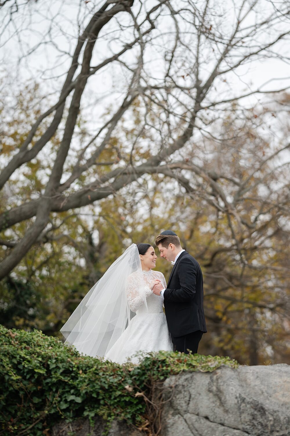 The couple pauses on a stone outcrop beneath bare branches, her veil catching the breeze.