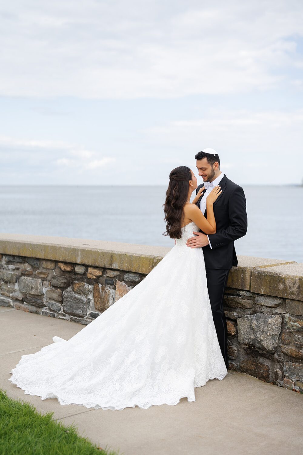 The couple stands at the stone seawall with the bride's train spread toward Long Island Sound.