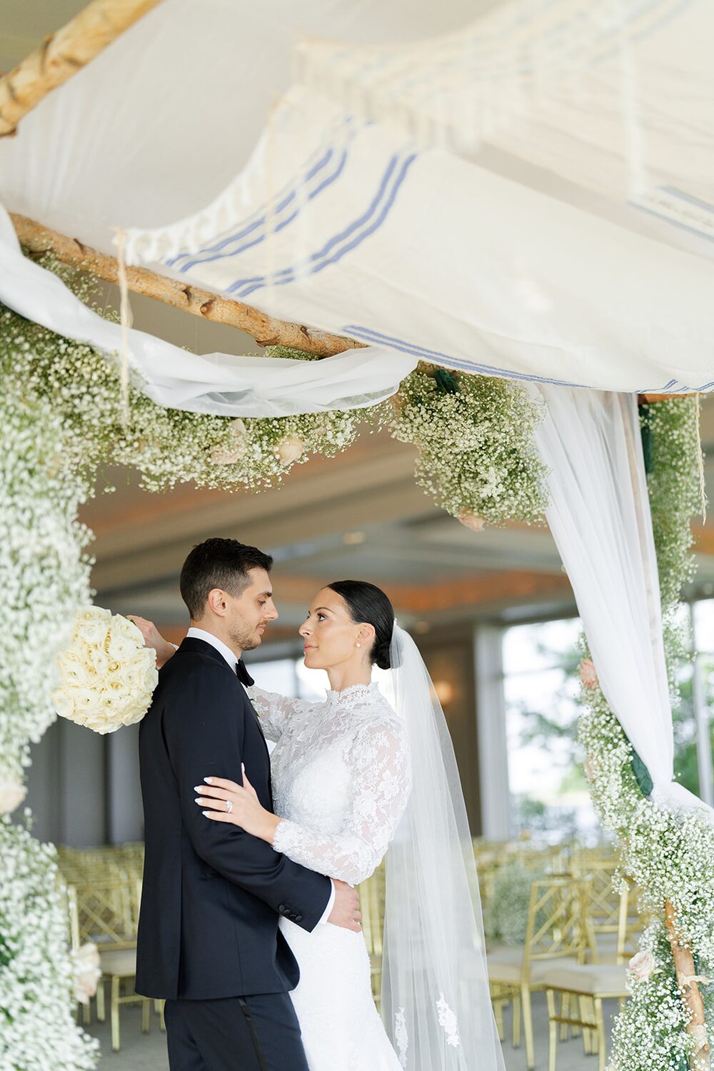 Under a tallit-draped chuppah wrapped in baby's breath, the couple stands together.