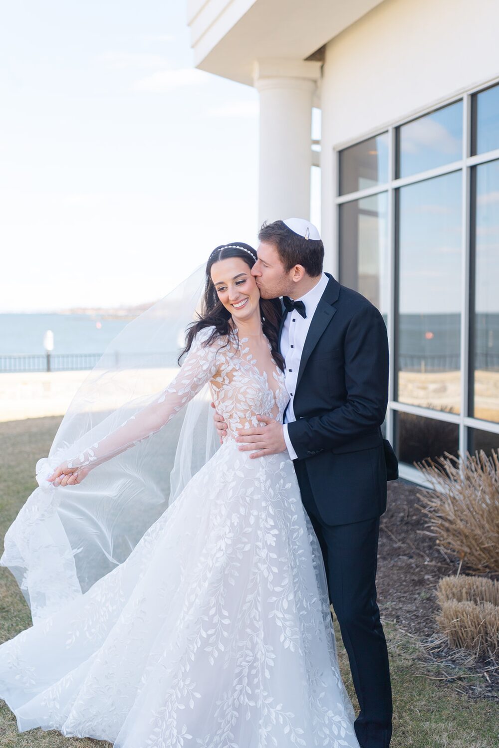 The groom kisses the bride's temple outside the venue's glass pavilion.