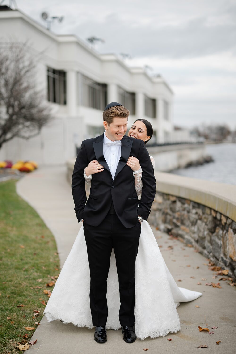 The bride wraps her arms around the groom on the waterfront path in soft November light.