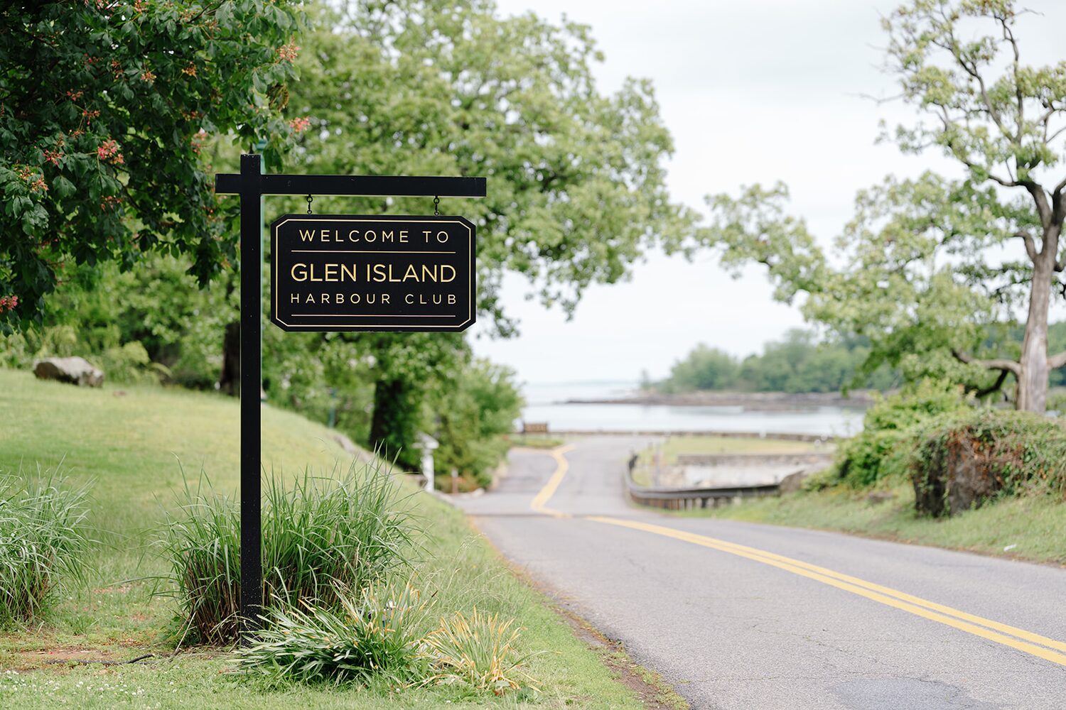 The Welcome to Glen Island Harbour Club sign at the park entrance, with the road curving toward the water.