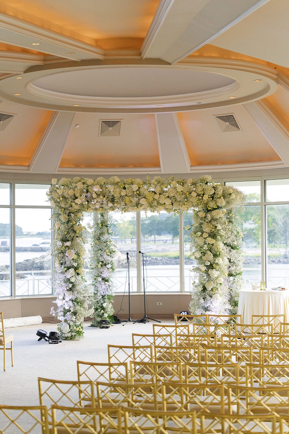 A white floral chuppah stands in the glass-walled ballroom with gold chairs and Long Island Sound beyond the windows.