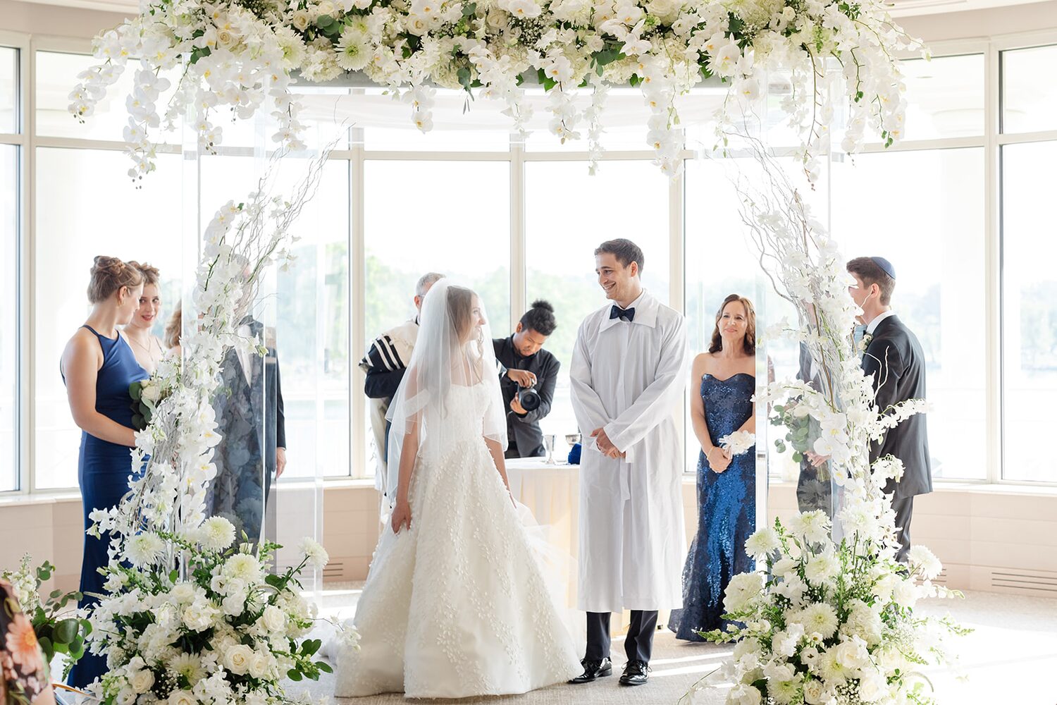 The groom in a white kittel faces his bride beneath the white floral chuppah.